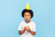 © khosrork - Portrait of cute little boy with curly hair wearing yellow party cone holding cake blowing candle making wish on his birthday. Indoor studio shot isolated on blue background.