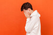 Portrait of depressed brunette young man wearing white hoodie hiding face in hand, looking desperate, is about to cry, empty copy space for text. Indoor studio shot isolated on orange background.