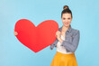 Portrait of romantic smiling woman with bun hairstyle wearing denim jacket holding big read heart saying about her feelings. Indoor studio shot isolated on light blue background.