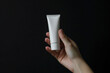 Hand gripping an empty white cosmetic cream tube against a matte black isolated solid background, highlighting elegance and premium skincare,