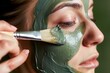 woman applying green clay mask on face with brush