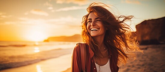 Wall Mural - A woman with long, layered hair is standing on a beach at sunset, smiling and looking happy. The water glistens beneath her as she enjoys the beautiful landscape