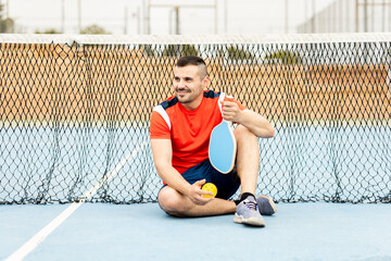 Wall Mural - The young man dressed in sportswear is sitting near the net of the court resting after training.An adult man between 30 and 40 years old is playing a game of pickleball outdoors.Pickleball concept.