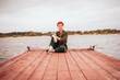 Portrait of a beautiful redhead woman sitting on a wooden deck or a pier by a river, looking straight to the camera