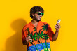 Photo of positive man with afro hairdo dressed print shirt in sunglass hold beer look at smartphone isolated on yellow color background