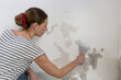 Saltpeter on the wall problem. Woman is using a scraper to scrape and remove all loose paint and plaster that is in poor condition, until a firm surface is achieved.	
