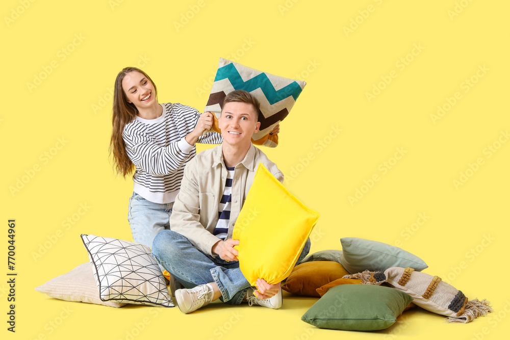 Young woman with her husband fighting pillows on yellow background