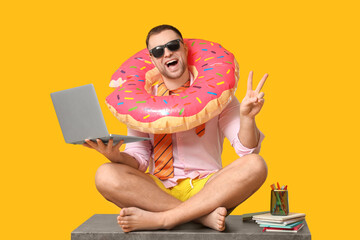 Wall Mural - Happy office worker in sunglasses with inflatable ring and laptop sitting on table and showing victory gesture against yellow background