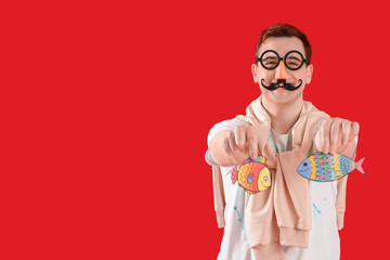 Wall Mural - Young man in funny disguise with paper fishes on red background. April Fool's Day celebration
