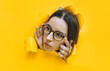 Portrait of a cute young woman with glasses looking through a hole in yellow paper. An incredulous look. Women's curiosity and gossip. Jealous wife.