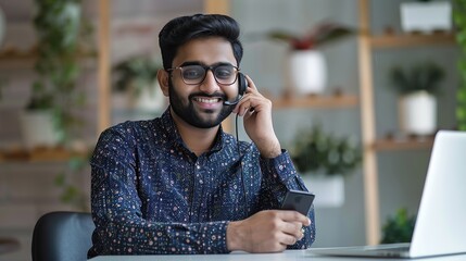 Poster - smiling young indian man freelancer talking on smartphone seated at desk with laptop, working at hom