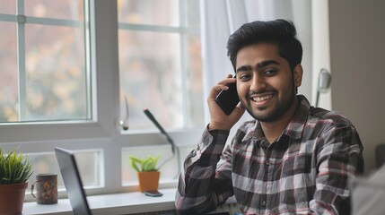 Poster - smiling young indian man freelancer talking on smartphone seated at desk with laptop, working at hom