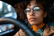 Young black woman driver with glasses behind the wheel, focused on the road ahead.
