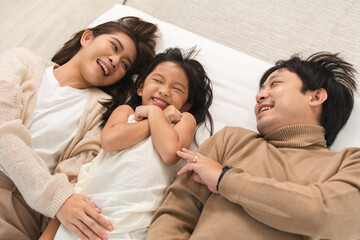 Wall Mural - Happy Asian family, father, mother, cute daughter having fun, lying on bed in bedroom at home, bond and relax together, smiling with happiness. Top view