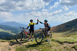Two cyclists men riding electric bikes outdoors. Male tourists resting on the top of hill, giving high five to each other, enjoying beautiful mountain landscape, wearing helmet and backpack.