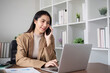 © wichayada - Young Asian business woman sits on the phone in an online business meeting using a laptop in a modern home office decorated with shady green plants.