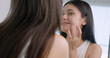 Beautiful young woman applies moisturizer to her face while standing in front of a mirror.