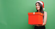 Cheerful young woman wearing a Santa hat holding a large gift box. Isolated on green background in studio.