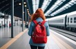 Young red-haired woman with a red backpack on her back waits at a large train station, rear view. 
Train travel. Railway station. Expectation