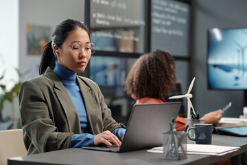 Aufkleber - Young serious businesswoman in formalwear looking at laptop screen during network while sitting by desk with windmill model
