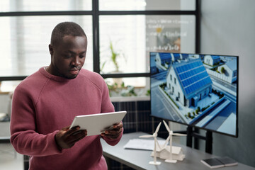 Leinwandbilder - African American male employee in casualwear networking or scrolling through project points against his workplace with interactive board