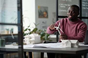 Aufkleber - Young smiling male architect rotating turbine of windmill model while sitting by workplace with unrolled blueprint and house mockup