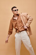 © LIGHTFIELD STUDIOS - Fashionable man in beige jacket on shoulder, shirt, pants and sunglasses on beige background, banner