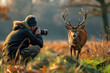 A wildlife photographer kneeling in the grass, holding his camera and taking pictures of a red deer running across him from behind