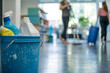 Detergents are placed in a bucket on the floor of an office, with two women cleaning in the background.