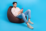 Fototapeta Panele - Full size photo of clever positive man wear sweatshirt denim pants sit on bean bag look at smartphone isolated on blue color background