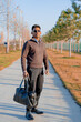 © oybekostanov - A man wearing a brown jacket and black pants is holding a black bag. He is standing on a sidewalk in front of trees