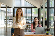 © Wasana - A confident businesswoman stands arms crossed in a modern collaborative workspace with a seated colleague in the background.