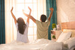 © WESTOCK - Back view shot of couples stretching hands after waking up from bed by seeing sunrise from window - concept of refreshment, morning beginning and peaceful harmony.
