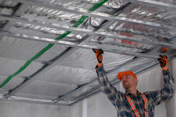 construction worker in overalls, gloves and tool belt assembles iron structure to lower plasterboard ceiling