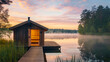 Wooden sauna house on the pier of a calm lake with mist and a colorful sunrise sky.