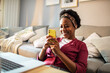 © Davor - Happy African American woman using smartphone on couch with laptop nearby