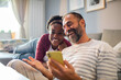 © Davor - Joyful interracial couple looking at smartphone and laughing together on sofa