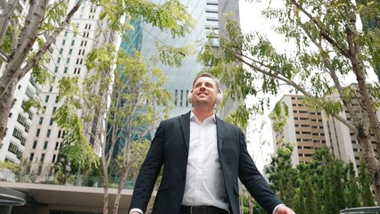 Wall Mural - Business people wear headphone to listen relaxing music while walking workplace at green city. Low angle camera of manager move to song while go to meeting surround with tree and skyscraper. Urbane.