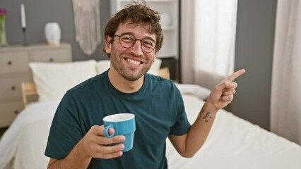 Wall Mural - Cheerful young hispanic man, sipping coffee in bed, giving a friendly point and smile, gleefully signaling to the side indoors. happy expression in cozy bedroom setting.