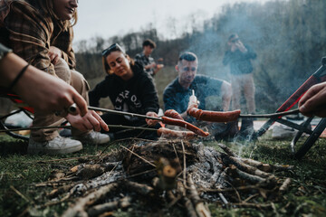 Wall Mural - A group of friends gather around a campfire, cooking sausages on sticks, enjoying nature and good company.