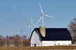A venerable white barn with an interior silo rests below towering wind turbines forming a contrast between traditional agriculture and the modern technology of wind farming. 