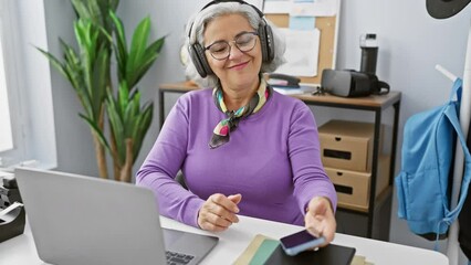 Sticker - A smiling mature woman with grey hair dances joyfully in an office environment while wearing headphones and using a smartphone.