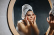 © qunica.com - A serene young woman performs a skincare routine, gently touching her face in front of a mirror in a cozy, dimly lit room.