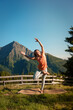 Yoga Pose Baum Variation - Sportliche Frau in Leggings und orangefarbenem Top macht Yoga mit Blick auf die Berge der Alpen unter strahlender Sonne