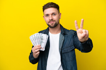 Wall Mural - Young caucasian man taking a lot of money  isolated on yellow background smiling and showing victory sign