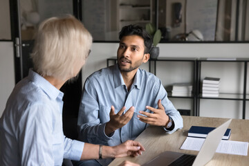 Canvas Print - Serious younger Indian professional man talking to elder colleague woman at workplace, discussing project, collaboration, management strategy. Business coworkers speaking at office table