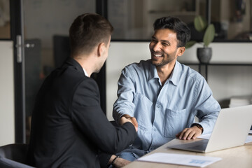 Wall Mural - Cheerful multiethnic male business partners shaking hands at workplace table, smiling, laughing, finishing conversation, negotiation, consultation meeting, discussing cooperation, partnership