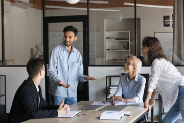 Wall Mural - Happy young Indian manager speaking to colleagues, offering creative ideas for business project. Diverse team of coworkers brainstorming at job meeting, enjoying communication