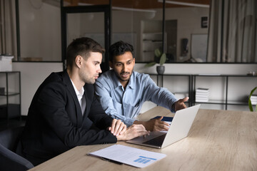 Serious multiethnic business colleagues working on job tasks together, talking at workplace table, watching online presentation on laptop, pointing hand at monitor, discussing creative ideas