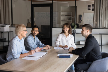 Wall Mural - Diverse small business team brainstorming on job tasks, planning cooperation, teamwork, discussing project, talking at meeting table. Senior manager talking speaking to younger colleagues